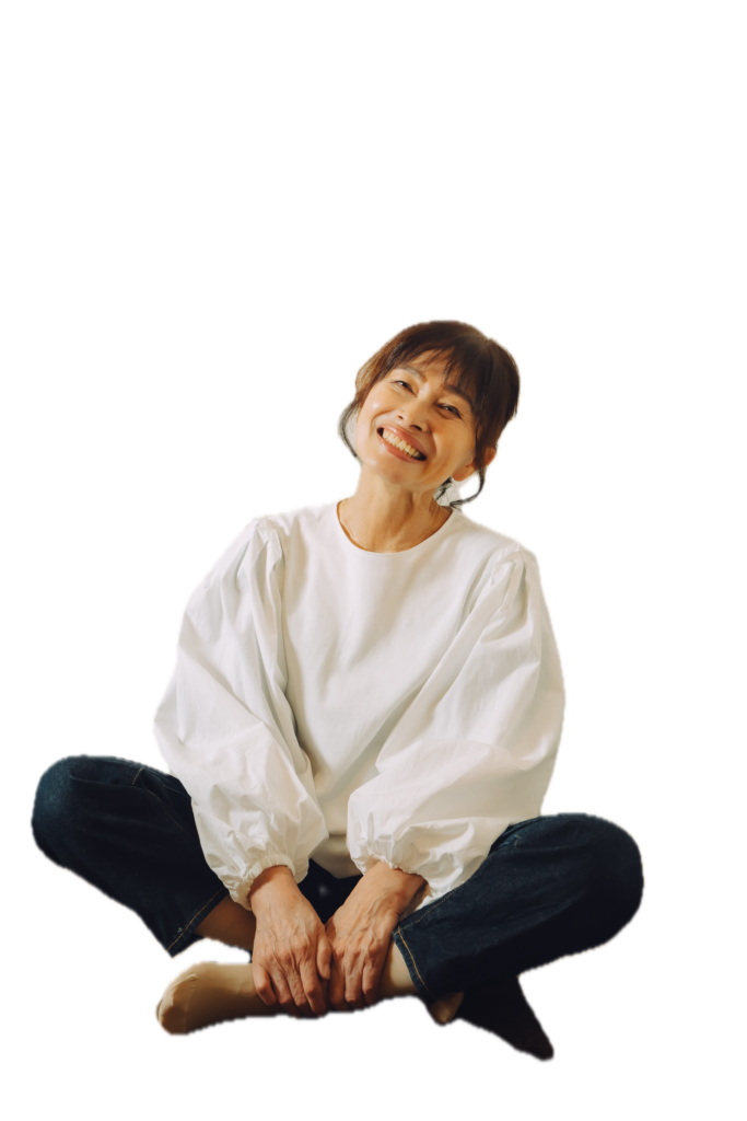 白いブラウスに紺色のパンツの女性。足を組んで少し顔を傾けやさ弾く微笑んでいる。リラックスしていて、好感度抜群。