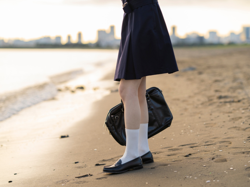 海岸で、女子高生が立っている。足元だけが写っているが、何か寂しげ